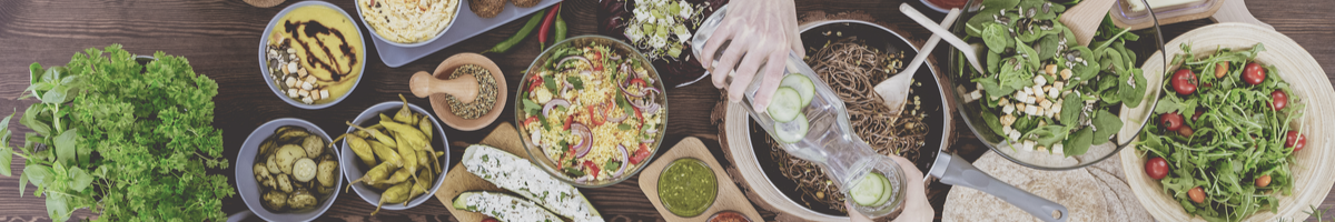 Auf einem Tisch stehen frische grüne Salate, gefüllte Zucchini, Antipasti, eine Pfanne mit gebratenen Vollkornspaghetti, Taboulé, Hummus und viele andere gesunde und leckere Speisen